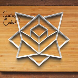 Puede incluir: Cortador de galletas geométrico blanco con diseño de estrella, con una forma de diamante central rodeada de formas triangulares y de diamante. El cortador está sobre una superficie de madera. El texto "Cactus Cookies" está en la esquina superior izquierda.