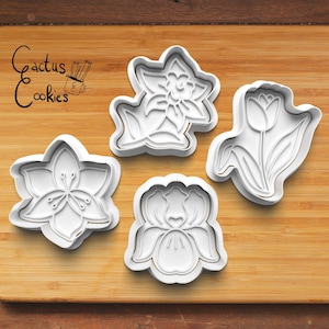 May include: Set of four white plastic cookie cutters shaped like flowers. The cutters are for making cookies in the shapes of a daffodil, a tulip, a cherry blossom, and an iris.