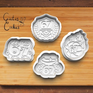 May include: Four white plastic cookie cutters with the number 67 in various designs. One cutter has the number 67 inside a shield with a laurel wreath. Another has the number 67 in flames. The other two cutters have the number 67 in a cartoon style. The text "Cactus Cookies" is in the upper left corner.