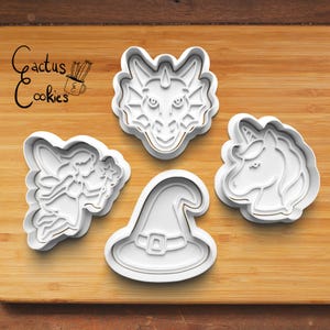 May include: Four white plastic cookie cutters in fantasy shapes: a dragon's head, a fairy, a witch's hat, and a unicorn. The cutters are on a wooden surface. The text "Cactus Cookies" is in the upper left corner.