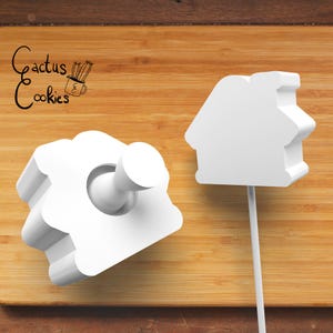 Puede incluir: Dos cortadores de galletas blancos con forma de casa sobre una superficie de madera. Uno tiene un mango y el otro está en un palo. El texto "Cactus Cookies" está en la esquina superior izquierda.