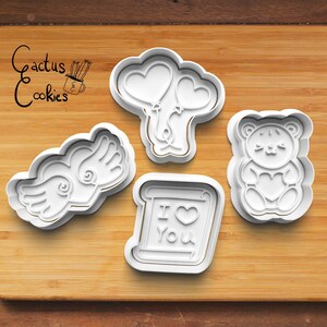 May include: Four white plastic cookie cutters on a wooden surface. The cutters are shaped like a heart with wings, a teddy bear holding a heart, a scroll with the text "I love you", and two balloons.