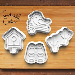 May include: Four white plastic cookie cutters in bird-watching theme. Includes a bird on a branch, a birdhouse, binoculars, and a sprig of leaves. The logo "Cactus Cookies" is in the upper left corner.