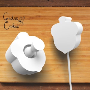 May include: Two white cookie cutters in the shape of an acorn and a flower. The acorn cutter is on a stick. The flower cutter has a cylindrical handle. The text "Cactus Cookies" is in the upper left corner.