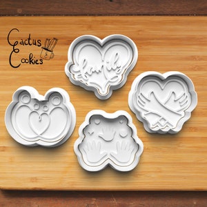 May include: Four white plastic cookie cutters on a wooden surface. The cutters are shaped like hearts, hands, and a family with a heart in the middle. The cutters are for making cookies.