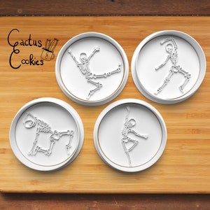 May include: Four white plastic cookie cutters shaped like skeletons in different dance poses. The cutters are on a wooden surface.