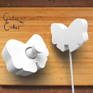 Può includere: Due tagliabiscotti in plastica bianca a forma di fiocco. Il fiocco a sinistra è piatto e il fiocco a destra è su un bastoncino.