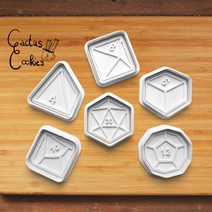 Puede incluir: Seis cortadores de galletas de plástico blanco con forma de dados diferentes, incluyendo un dado de cuatro caras, un dado de seis caras, un dado de ocho caras, un dado de diez caras, un dado de doce caras y un dado de veinte caras. Los cortadores están sobre una superficie de madera.