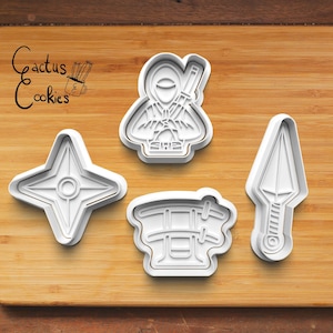 Puede incluir: Cuatro cortadores de galletas de plástico blanco con forma de ninja, shuriken, katana y templo japonés. Los cortadores de galletas están sobre una superficie de madera.