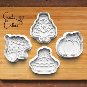 Może przedstawiać: Cztery białe plastikowe wykrawacze do ciastek w kształcie indyka, dyni, ciasta i żołędzia z liśćmi. Wykrawacze do ciastek znajdują się na drewnianej powierzchni. Tekst "Cactus Cookies" znajduje się w lewym górnym rogu.