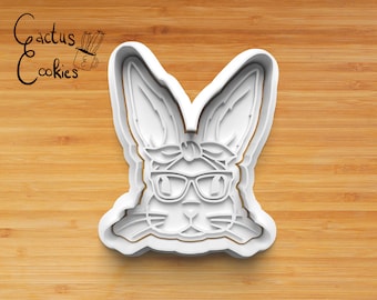 May include: White plastic cookie cutter shaped like a bunny wearing sunglasses and a bandana. The bunny has large ears, a small nose, and a cute expression.