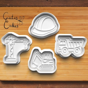 May include: Four white plastic cookie cutters shaped like construction vehicles and equipment. The shapes include a hard hat, a crane, an excavator, and a dump truck. The words "Cactus Cookies" are in the upper left corner.