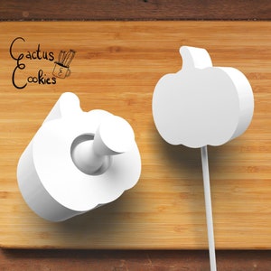 May include: Two white pumpkin-shaped cookie cutters on a wooden surface. The cutter on the left is lying flat, while the cutter on the right is standing upright on a stick.