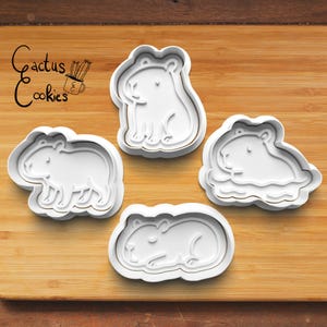 May include: Four white plastic cookie cutters in the shape of capybaras on a wooden surface. The capybaras are in different poses, including sitting, standing, and lying down. The text "Cactus Cookies" is in the upper left corner.