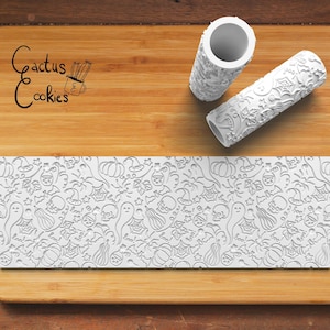 May include: Two white rolling pins with a Halloween themed design. The design includes ghosts, pumpkins, bats, spiders, and stars. The rolling pins are on a wooden surface with the text "Cactus Cookies" in black.
