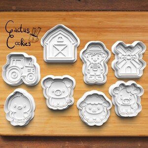 May include: A collection of white plastic cookie cutters in various farm-themed shapes, including a tractor, barn, farmer, windmill, chick, pig, sheep, and cow. The words "Cactus Cookies" are in the upper left corner.