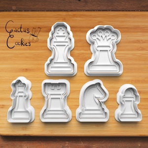 May include: Set of six white plastic cookie cutters shaped like chess pieces: king, queen, rook, knight, bishop, and pawn. The cutters are on a wooden surface.