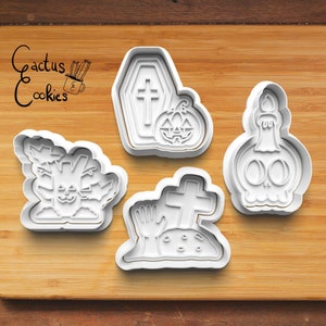 Może przedstawiać: Białe plastikowe wykrawacze do ciastek o tematyce Halloween na drewnianej powierzchni. Wykrawacze obejmują trumnę z krzyżem, dynię, butelkę w kształcie czaszki ze świecą, drzewo z twarzą i nagrobek z ręką. Tekst "Cactus Cookies" znajduje się w lewym górnym rogu.