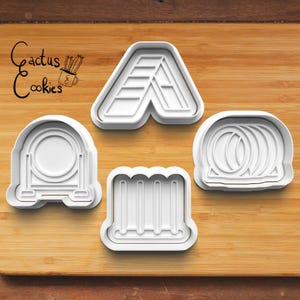 May include: Four white plastic cookie cutters in various shapes, including the letter "A", a circle, a series of vertical lines, and a series of concentric circles. The cutters are on a wooden cutting board. The words "Cactus Cookies" are in the upper left corner.