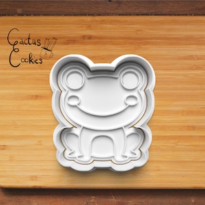 Op de afbeelding: Witte plastic koekjesvormer in de vorm van een cartoon kikker met een brede glimlach en grote ogen. De vormer ligt op een houten oppervlak.