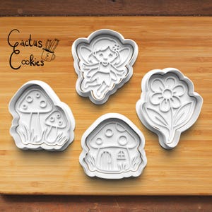 Op de afbeelding: Vier witte plastic koekjessnijders in de vorm van een fee, een bloem, een paddenstoel en een paddenstoelhuis. De uitstekers liggen op een houten oppervlak. De tekst "Cactus Cookies" staat in de linkerbovenhoek.