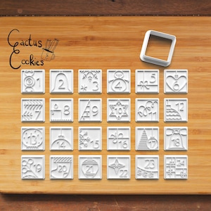 May include: A set of 24 white plastic cookie cutters shaped like Christmas-themed designs, including numbers 1 through 24, snowflakes, stars, bells, and other holiday symbols. The cutters are arranged in four rows of six on a wooden surface.