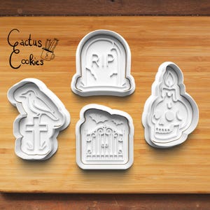 Może przedstawiać: Cztery białe plastikowe wykrawacze do ciastek o tematyce Halloween na drewnianej powierzchni. Wykrawacze obejmują nagrobek z napisem "RIP", kruka na krzyżu, czaszkę ze świecą i bramę z nietoperzami. Logo "Cactus Cookies" znajduje się w lewym górnym rogu.
