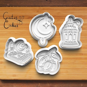 May include: Four white plastic cookie cutters shaped like a crescent moon and star, a mosque with a crescent moon, a lantern, and a branch of olives. The cookie cutters are on a wooden surface. The text "Cactus Cookies" is in the top left corner of the image.