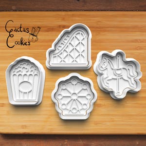 Könnte beinhalten: Vier weiße Ausstecher aus Kunststoff in Form eines Popcornbehälters, einer Achterbahn, eines Riesenrads und eines Karussellpferds. Der Text "Cactus Cookies" befindet sich in der oberen linken Ecke. Die Ausstecher befinden sich auf einer Holzoberfläche.