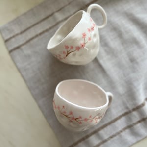 Puede incluir: Dos tazas de cerámica blanca con diseños florales rosas. Las tazas tienen una superficie texturizada y una forma redondeada.