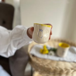 Peut inclure: Une tasse en céramique blanche avec un bord jaune et un dessin de citron bleu dessiné à la main. La tasse est tenue par une main.