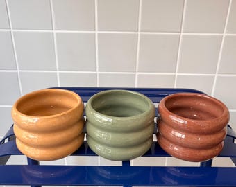 Puede incluir: Tres macetas de cerámica con un diseño ondulado y acanalado. Las macetas son en tonos de amarillo, verde y marrón.
