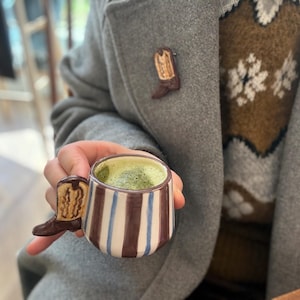 May include: A ceramic mug with a cowboy boot handle and a brown and blue striped design. The mug is filled with a green beverage. A cowboy boot pin is attached to a grey coat.