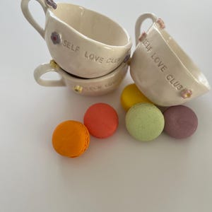 May include: Four white ceramic teacups with the words "Self Love Club" on the side. The cups are stacked on top of each other. There are five colorful macarons in the background.