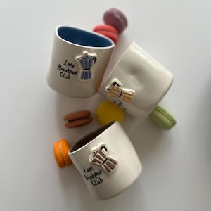 Puede incluir: Cuatro tazas de cerámica blanca con un diseño de cafetera azul, marrón y amarillo. Cada taza tiene el texto "Late Breakfast Club" escrito en ella.