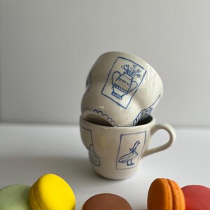 May include: Two white ceramic mugs with blue hand-painted designs. The top mug features a vase with flowers, while the bottom mug has a bird design. Four colourful macarons are in the foreground.