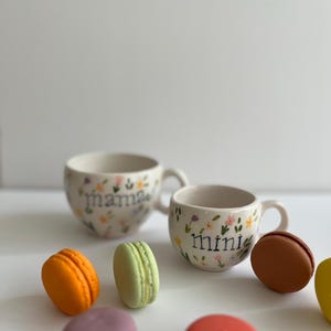 May include: Two white ceramic mugs with floral designs. The larger mug says "mama" and the smaller mug says "mini".  Five colorful macarons are scattered around the mugs.