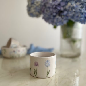 May include: White ceramic planter with a blue and purple floral design. The planter is round and has a smooth surface.