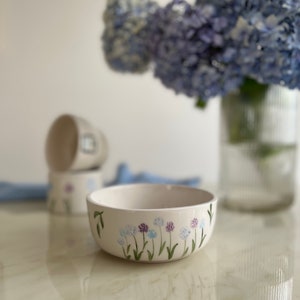 Peut inclure: Bol en céramique blanche avec un motif floral composé de fleurs bleues, violettes et vertes. Le bol est posé sur une surface blanche.