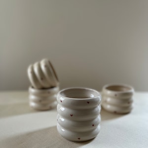 May include: Four white ceramic cups with a textured, ribbed design and small red hearts. The cups are arranged on a white surface.