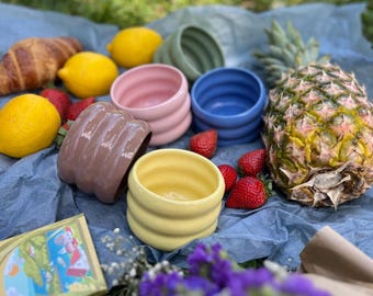 Puede incluir: Una escena de picnic con cuencos de cerámica coloridos, fruta fresca y un cruasán. Los cuencos son en tonos rosa, azul, amarillo, verde y marrón. También hay fresas, limones y una piña.