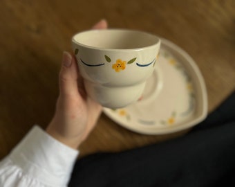 May include: A white ceramic teacup with a blue and yellow floral design. The cup is being held in a hand and is sitting on a white saucer.