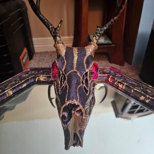 May include: A decorative deer skull with a purple and gold glitter design. The skull features gold accents, pink gems in the eye sockets, and dark brown antlers with a leopard print pattern. The skull is reflected in a mirror.