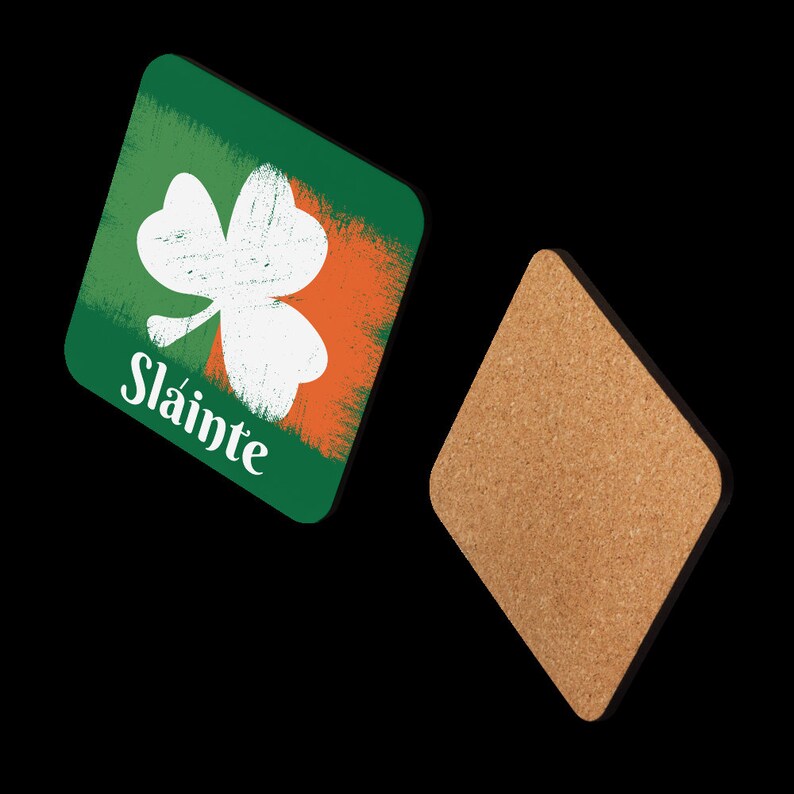 Puede incluir: Un posavasos cuadrado de corcho con un dise&ntilde;o de tr&eacute;bol verde, blanco y naranja y la palabra "Sl&aacute;inte" impresa en &eacute;l. El posavasos est&aacute; hecho de corcho y tiene un borde negro.