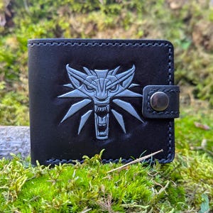 Puede incluir: Cartera de cuero negro con un emblema de lobo plateado. La cartera plegable tiene un cierre de botón a presión y bordes cosidos. El emblema del lobo está grabado en la parte delantera. La cartera está sobre musgo verde.