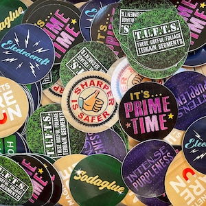 Puede incluir: Una colección de pegatinas redondas de colores con varios diseños y textos. Las pegatinas presentan frases como "It's Prime Time", "T.U.F.T.S. These Useful Foliage Terrain Segments", y "Sharper Is Safer".