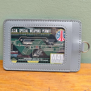 Może przedstawiać: Szary uchwyt na karty z przezroczystym oknem, na którym widnieje zielony schemat broni, oznaczony jako "M41 Pulse Rifle". Tekst zawiera "S.C.M. Special Weapons Permit" i "U.S. Colonial Marine Personnel Database."