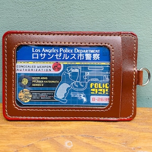 Könnte beinhalten: Ein brauner Lederkartenhalter mit roter Umrandung, der eine blaue Karte mit dem Text "Los Angeles Police Department" und "Concealed Weapon Authorization" zeigt. Der Halter hat einen Metallring.