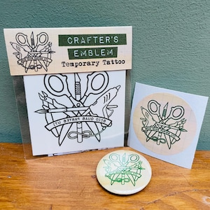 May include: A temporary tattoo set featuring a crafter's emblem design with scissors, a glue gun, and a needle. The set includes a temporary tattoo, a sticker, and a button. The text "CRAFTER'S EMBLEM" is displayed on the packaging.