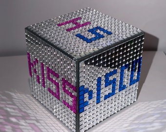 Puede incluir: Un cubo espejado con la palabra "MSU" escrita en mosaicos azules y rosas. El cubo está cubierto de pequeños cuadrados reflectantes, creando un efecto brillante. El cubo está sobre una superficie blanca.